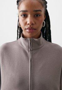 Top zíper taupe claro com gola alta e textura suave. Apresenta um design simples, sem padrões ou acentos proeminentes.