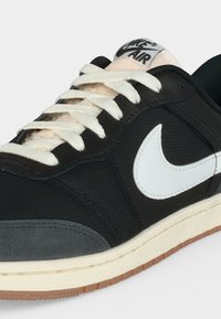 Μαύρο και γκρι αθλητικό παπούτσι Nike Air με λευκά κορδόνια και μεγάλο λευκό λογότυπο Nike στο πλάι, μπεζ σόλα με ορατό καφέ πέλμα.