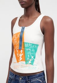 Femme portant un haut blanc sans manches avec des imprimés graphiques orange et verts, une fermeture éclair bleue verticale et un jean avec une étiquette « Replay ».