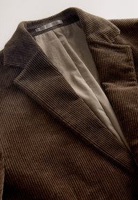 Blazer en velours côtelé marron avec une texture côtelée distincte, un col  à revers cranté et une doublure beige contrastante visible à l'intérieur.