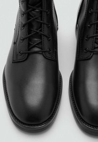 Botas de cuero negro con una textura suave, que presentan un diseño de cordones, punta redondeada y costuras en contraste a lo largo de la suela.