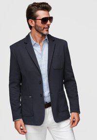 Homme portant un blazer sombre, une chemise rayée bleu clair, un pantalon blanc, des lunettes de soleil, regardant sur le côté sur un fond uni.