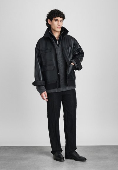 Giovane uomo che indossa una giacca di pelle nera oversize, un maglione di lana grigio, pantaloni neri texturizzati e scarpe eleganti nere, in piedi davanti a un muro semplice.
