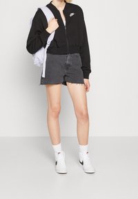 Černý zkrácený zipový svetr, tmavé džíny s ošoupaným lemem, bílé vysoké tenisky a světle fialový batoh.