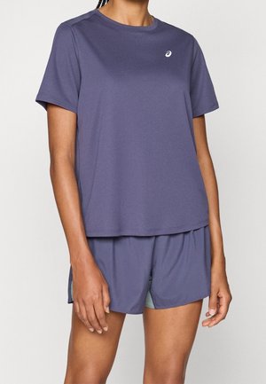 Frau mit lila kurzärmligem Sportshirt und passenden Shorts mit grünem Innenfutter vor einfarbigem Hintergrund.