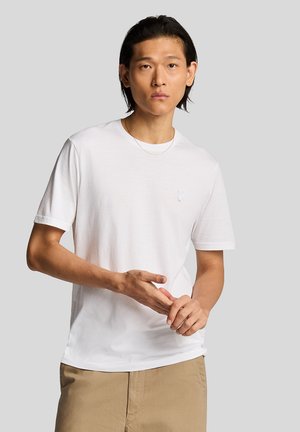 Junger Mann mit schwarzem Haar, der ein weißes T-Shirt und beige Hosen trägt, passt seine Finger mit neutralem Gesichtsausdruck vor einem schlichten Hintergrund an.