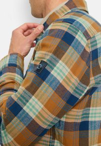 Kariertes Hemd in braunen, blauen und grünen Tönen; verfügt über einen strukturierten Stoff, einen Knopfkragen und ein Logopatch an der linken Schulter.