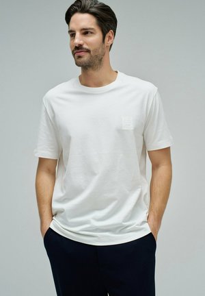 Hombre con cabello oscuro y barba, vistiendo una camiseta blanca de manga corta lisa y pantalones negros, con las manos en los bolsillos, mirando hacia un lado.