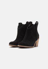 Botines de ante negro con un tacón de madera maciza, con detalles trenzados a los lados y tiradores para poner y quitar fácilmente.