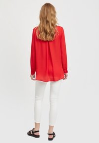 Blusa rossa a maniche lunghe con una vestibilità ampia, tessuto morbido e polsini con bottoni, abbinata a pantaloni bianchi slim-fit e sandali neri.
