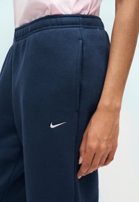 Γυναίκα που φοράει σκούρο μπλε φόρμα Nike με λευκό λογότυπο στο αριστερό μηρό, στέκεται με το δεξί χέρι χαλαρό δίπλα στο σώμα.