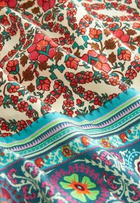 Tissu avec des motifs floraux et botaniques éclatants en rouge, marron, sarcelle et vert sur fond crème, bordé de motifs géométriques complexes turquoise et rose.
