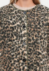 Γυναίκα που φοράει ένα κουμπωμένο, φανελένιο μπουφάν με πράσινο, μπεζ και μαύρο λεοπάρ σχέδιο πάνω από ένα ανοιχτό ροζ πουλόβερ.