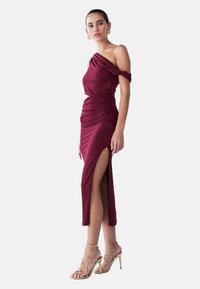 Burgundy off-shoulder dress with ruched detailing, knee-high slit, and soft texture, paired with strappy gold high heels.