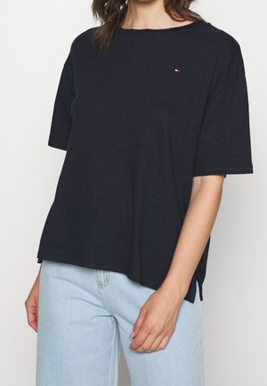 Kvinde iført en løs, sort kortærmet skjorte med et lille logo og lyseblå jeans, stående mod en ensfarvet baggrund.