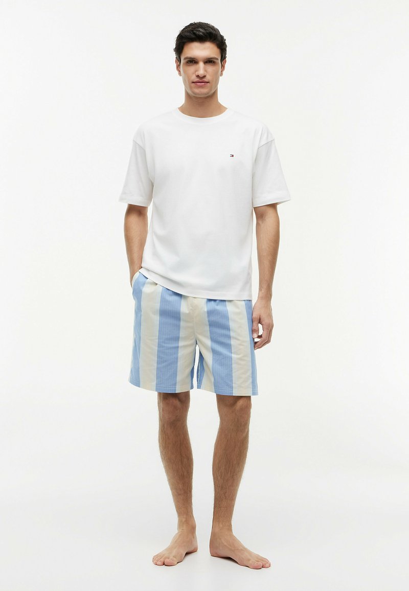 Homme debout, pieds nus, portant un t-shirt blanc à manches courtes et un short à rayures verticales bleues et beiges, sur un fond uni.