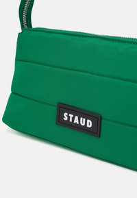Zielona torba z pikowanej nylonu z srebnym zamkiem błyskawicznym oraz prostokątną czarną metką z napisem "STAUD" w białym kolorze.