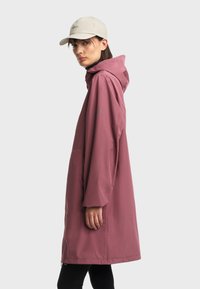 Žena u ljubičastom kišnom plaštu s kapuljačom i bež šeširom, stoji u profilu protiv jednostavne svijetlosive pozadine.