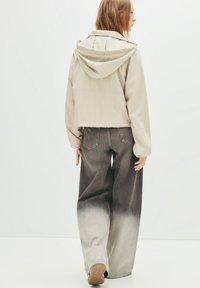 Beige Jacke mit Kapuze, kurze Form; kombiniert mit weiten Jeans, die einen Farbverlauf von grau zu hell zeigen. Glatte Stoffoberfläche.
