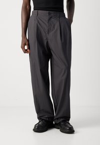 Man wearing loose dark gray trousers and black leather shoes, standing with one hand in pocket against plain white background.