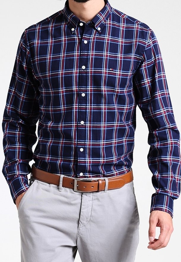 Camisa de cuadros azul marino con acentos en rojo y blanco, cuello abotonado y mangas largas. Combinada con un cinturón marrón y pantalones gris claro.