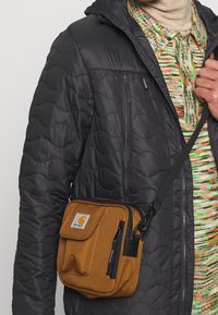 Borsa a tracolla in tessuto marrone con zip superiore e tasca frontale, dotata di un'etichetta con logo. Indossata sopra una giacca nera isolante.