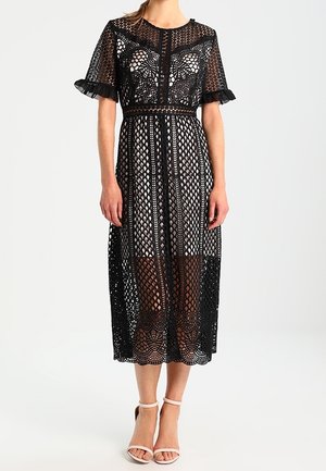 Robe midi noire en dentelle avec un motif géométrique, manches courtes, taille cintrée et accents froncés aux manches. Portée avec des talons à lanières.