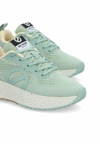 Baskets vert clair avec un dessus texturé, une semelle blanche mouchetée, et un logo bien visible sur la languette. Comprend des lacets plats pour le maintien.