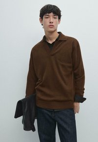 Brauner Strickpullover mit V-Ausschnitt, geripptem Saum und langen Ärmeln, getragen über einem schwarzen Hemd mit Kragen. Das Modell hält eine schwarze Lederjacke.