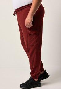 Bordeauxfarbene Sweatpants mit einem Kordelzugbund, Cargo-Tasche und gerippten Bündchen, kombiniert mit schwarzen Sportschuhen. Glatte Stofftextur.