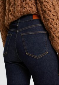 Jeans de mezclilla azul oscuro con un diseño ajustado, costuras amarillas en contraste y dos bolsillos traseros. Combinados con un suéter marrón texturizado.