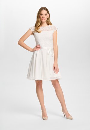 Jeune femme dans une courte robe blanche en dentelle avec manches courtes et escarpins beige, debout avec la main sur la hanche devant un fond uni.