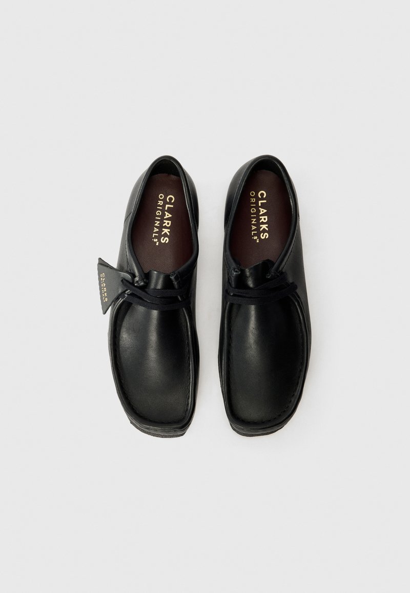 Svarta läderskor med rund tå, minimal sömnad, elastiska snören och en texturerad gummisula. Har en tagg med "CLARKS ORIGINALS."