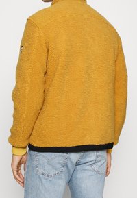 Giacca in pile giallo senape con superficie texturizzata, orlo nero e colletto alto. Presenta polsini a coste e un logo sulla manica.