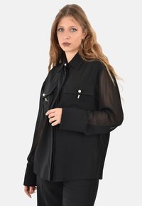 Camicia nera con bottoni, maniche lunghe trasparenti, due tasche frontali e bottoni simili a perle. Taglio dritto e tessuto liscio.