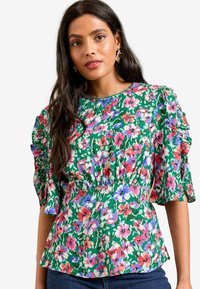 Ziedu blūze ar zaļu fonu, kurā redzami rozā, purpura un baltie ziedi. Īsas volānu piedurknes un elastīgs jostasvieta.