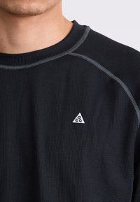Schwarzes, strukturiertes Langarmshirt mit rundem Ausschnitt und grauen Stichdetails. Verfügt über ein kleines weißes, dreieckiges Logo auf der Brust.