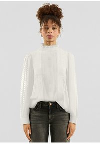 Witte blouse met een hoge gekruiste kraag, gestructureerde stof met plooitjes en gestippelde patronen, lange mouwen, gecombineerd met donkere spijkerbroek.