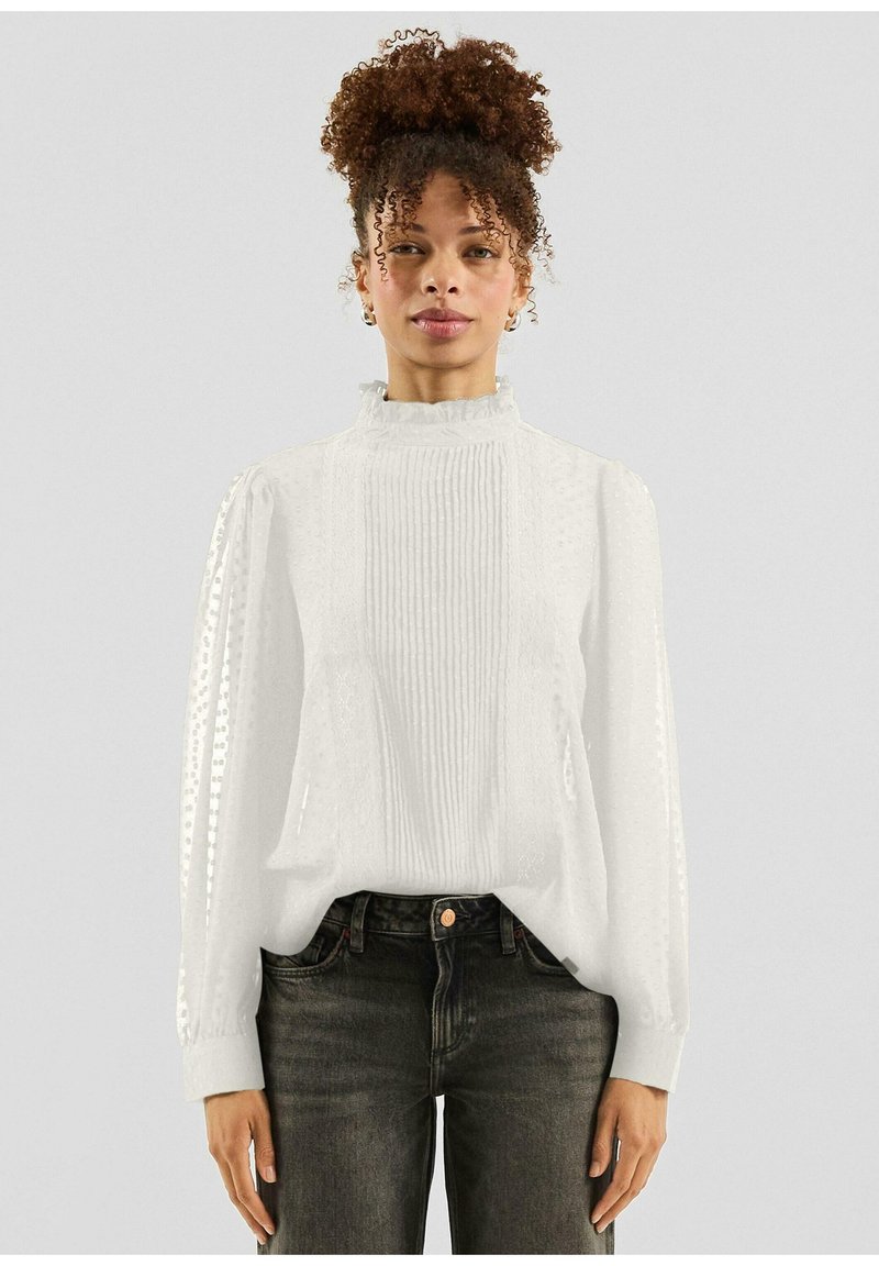 Witte blouse met een hoge gekruiste kraag, gestructureerde stof met plooitjes en gestippelde patronen, lange mouwen, gecombineerd met donkere spijkerbroek.