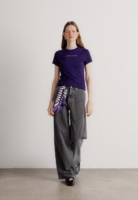 Violeta kokvilnas T-kreklu ar logotipu, kas apvienots ar pelēkām plašajām biksēm. Strīpains violets un balts šalle, kas sasieta ap jostasvietu, piešķir vizuālu interesi.