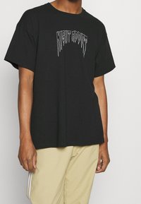 Černé bavlněné tričko s krátkým rukávem, které na hrudi zdobí bílé stylizované textové grafické znázornění "NIGHT ADDICT".