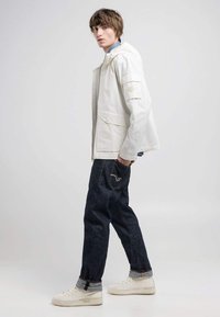 Chaqueta blanca con cremallera y capucha, combinada con jeans de mezclilla oscura y zapatillas blancas. El conjunto presenta un ajuste relajado y casual.