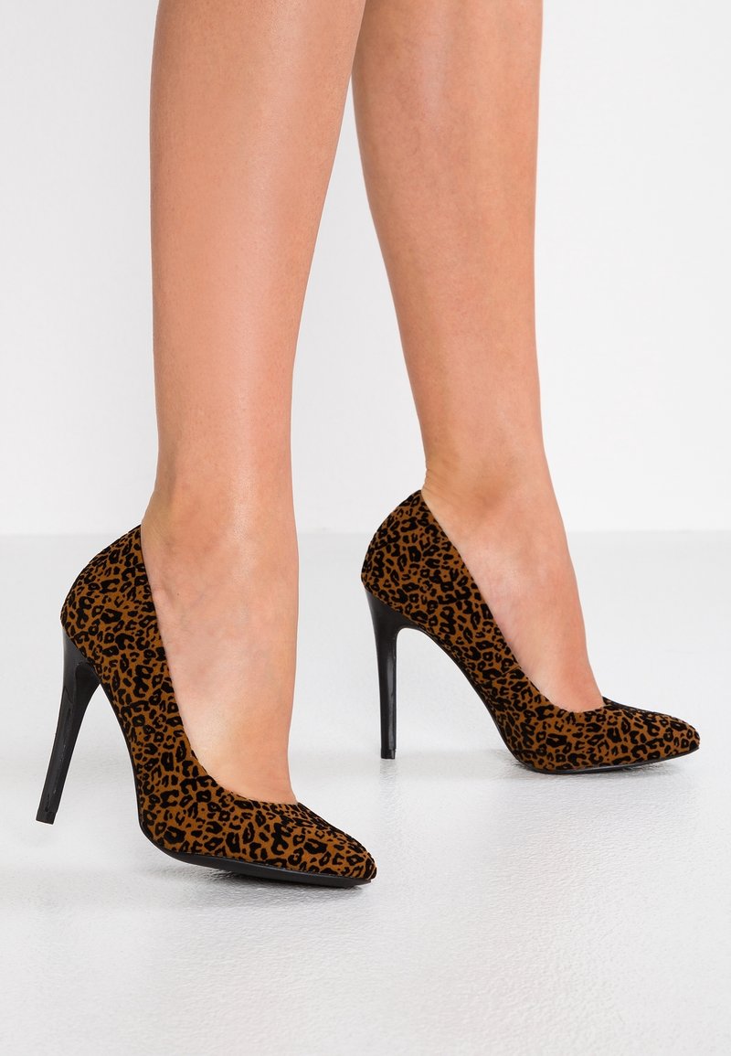 Jambes féminines portant des escarpins à talons hauts imprimé léopard marron avec des bouts pointus sur un sol blanc et un mur en arrière-plan.