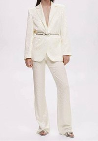 Biały, teksturowany garnitur z głębokim dekoltem w serek, ze metalowym paskiem, w połączeniu z rozwijającymi się spodniami. Gładki materiał z subtelnymi detalami wzoru.