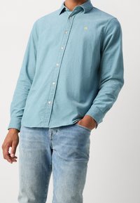 Camicia azzurra chiaro a maniche lunghe con bottoni, realizzata in materiale strutturato, caratterizzata da un sottile logo sul petto e abbinata a jeans in denim chiaro.