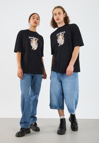 Sorte grafiske t-skjorter med teksten "Unreleased" og fargerik kunst, kombinert med lyseblå denimjeans og svarte støvler.