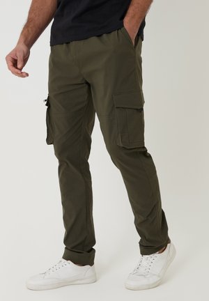 Mann trägt olivgrüne Cargohosen mit großen Seitentaschen und weißen Sneakers, steht mit einer Hand in der Tasche.