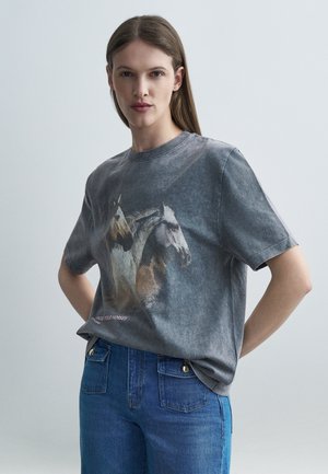 Femeie cu păr lung purtând un tricou gri spălat, pe care sunt imprimate doi cai, asortat cu blugi albastri cu talie înaltă, pozând pe un fundal simplu.