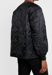 Musta tikattu takki timantinmuotoisella kuviolla, valmistettu kiiltävästä nylonmateriaalista. Takissa on resorit hihansuuissa ja rento istuvuus.