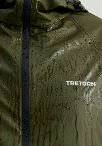 Vattentät olivgrön jacka med svart dragkedja och huva. Har vattendroppar på ytan och en vit logotyp. Slät textur.